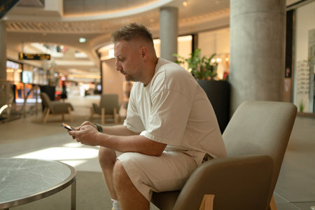 Handsome Caucasian adult man in stylish casual clothes using mobile phone on a soft chair at shopping mallの写真素材