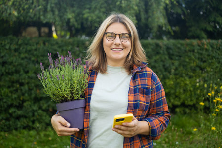 Gardening, greenery backyard garden concept. Young happy female gardener holding lavender plant and used smart phone in backyard garden. High quality photoの写真素材
