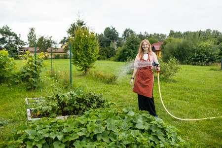 30s woman watering fresh plants growing at home vegetable garden. Gardener taking care of plants at the backyard of her house. Concept of sustainability and growing organic. High quality photoの写真素材