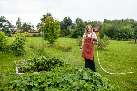 30s woman watering fresh plants growing at home vegetable garden. Gardener taking care of plants at the backyard of her house. Concept of sustainability and growing organic. High quality photoの写真素材