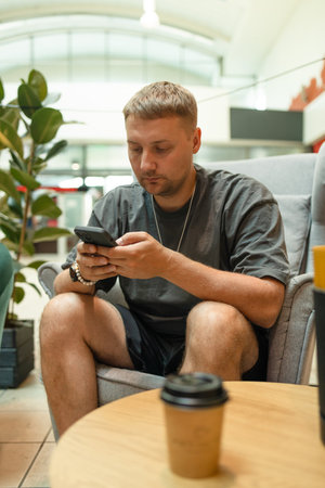 Young man with phone and power bank. Takeaway paper coffee cup with aroma latte coffee on a wood table at city cafe. Male with social media addiction on smartphone scrolling internet,の写真素材