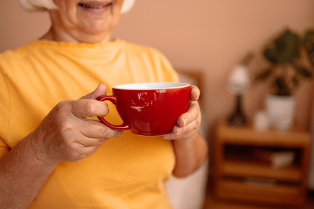 Happy Caucasian blonde senior woman sitting on a white bed with a cup in her hand. Female relaxing and drinking cup of hot coffee or tea in the bedroom.の写真素材