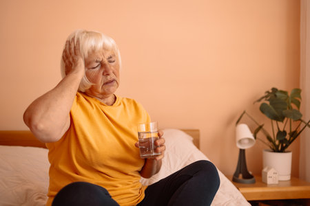 Senior 60s woman in pajamas suffering from headache sitting on bed at home bedroom, holding a glass of water, health, old age and people conceptの写真素材