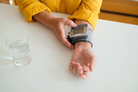 Healthy woman self checking blood pressure using machine. Senior woman with hypertension measuring blood pressure herself at home. Heart rate measurement. Glass of water on the tableの写真素材