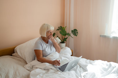Beautiful senior 70s woman yawns as wants to sleep, sad to wake up early in morning using computer in bed. 60s female watching video on laptop in her bedroom, e-learning or browsing internet websites, online shopping at home.の写真素材
