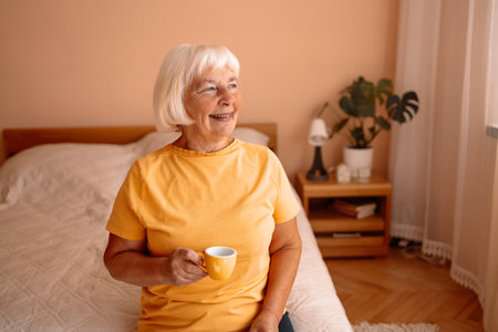 Happy Caucasian blonde senior woman sitting on a white bed with a cup in her hand. Female relaxing and drinking cup of hot coffee or tea in the bedroom.の写真素材