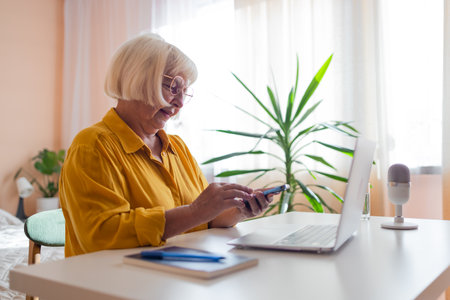 Focused old woman at home using laptop. Senior stylish entrepreneur with notebook and pen wearing eyeglasses working on computer. Serious lady analyzing and managing domestic bills and home finance. High quality photoの写真素材