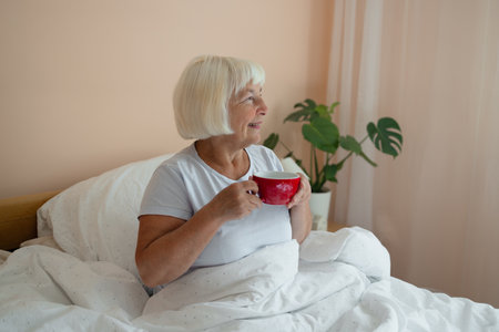 Relaxed Caucasian senior blonde hair woman in white pajama sitting on bed and drinking latte coffee on bed in weekend. High quality photoの写真素材