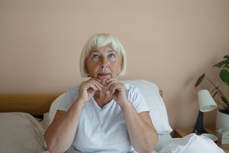 Close up of smiling Caucasian 50s senior woman siting on bed and applying face cream. Woman doing beauty care face massage at home.の写真素材