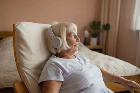 Elderly 60s 70s woman in headphones lying on chair and listening to music with closed eyes while resting in cozy living room at homeの写真素材