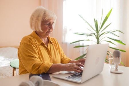 Focused old woman at home using laptop. Senior stylish entrepreneur with notebook and pen wearing eyeglasses working on computer. Serious lady analyzing and managing domestic bills and home finance. High quality photoの写真素材