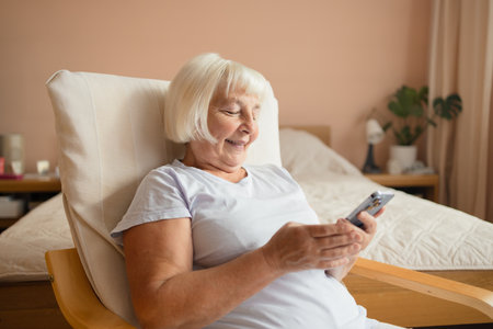 Caucasian 60s 70s happy woman using cell phone relaxing at home. Smiling senior lady holding smartphone enjoying mobile shopping, checking social media messages, playing game or buying online.の写真素材
