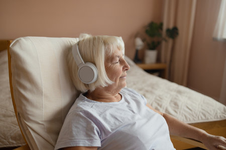 Elderly 60s 70s woman in headphones lying on chair and listening to music with closed eyes while resting in cozy living room at homeの写真素材