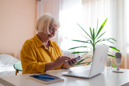 Focused old woman at home using laptop. Senior stylish entrepreneur with notebook and pen wearing eyeglasses working on computer. Serious lady analyzing and managing domestic bills and home finance. High quality photoの写真素材