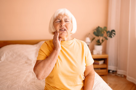 Unhappy sad Caucasian 60s 70s senior woman with toothache sits on a bed in the living room. Female pensioner in yellow shirt holding hand on her face having worried uneasy facial expression, thinking about her future.の写真素材