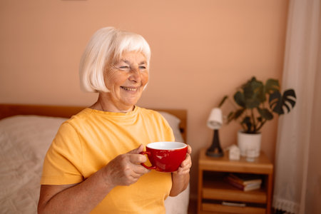 Happy Caucasian blonde senior woman sitting on a white bed with a cup in her hand. Female relaxing and drinking cup of hot coffee or tea in the bedroom.の写真素材