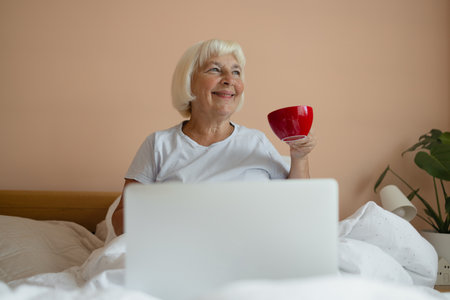 Caucasian senior 60s woman sitting with cup of coffee on bed and having breakfast while enjoying morning in bedroom at home. Female using laptop. Morning rituals.の写真素材