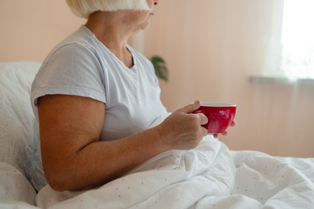 Relaxed Caucasian senior blonde hair woman in white pajama sitting on bed and drinking latte coffee on bed in weekend. High quality photoの写真素材