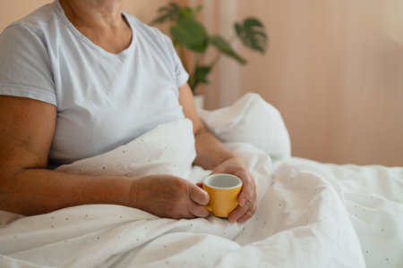 Relaxed Caucasian senior blonde hair woman in white pajama sitting on bed and drinking coffee on bed in weekend. High quality photoの写真素材