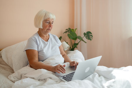Happy casual beautiful senior 60s woman working on a laptop sitting on the bed in the house. Freelancer, distant work, work at home, online education. Self isolation.の写真素材