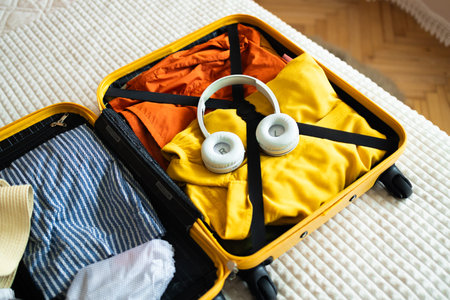 Open yellow suitcase with different family clothes and accessories on bed in light bedroom....