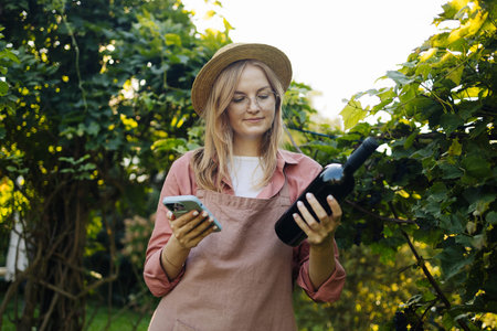 Caucasian blonde woman vintner relaxing in the vineyards in summer season. Girl in hat and an apron...