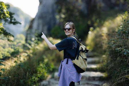 Photo of a young smiling woman carrying a backpack and hiking in the nature.Portrait of a happy 30s hiker female standing on the slope of mountain ridge against mountains. woman is happy and looking at camera. Ojcowski parkの写真素材