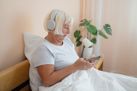 Elderly woman wearing headphones lying on bed using smartphone, embracing technology and enjoying a peaceful moment in her bedroom.の写真素材