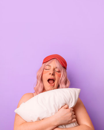 Sleeping 20s 30s woman in home wear having fun while resting wear sleep mask yawns as wants to sleep, embraces white soft pillow against purple background.の写真素材