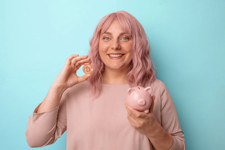Young caucasian woman smiling holding a piggybank and bitcoin for investment on light blue backgroundの写真素材