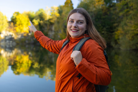 Happy female with backpack on hike in nature. Beautiful woman hiker standing on mountain trail near a lake and looking at camera smiling shows a direction. Lifestyle, adventure, nature, active life. High quality photoの写真素材