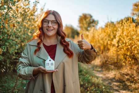Happy smiling woman holding a mock up of a house in her hands while standing outdoors at an autumn park. High quality photoの写真素材