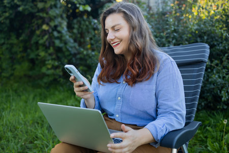 Close up portrait of smiling female gardener sitting on chair in garden, using laptop phone and speaking on web call surrounded by exotic plants. Home gardening, freelanceの写真素材