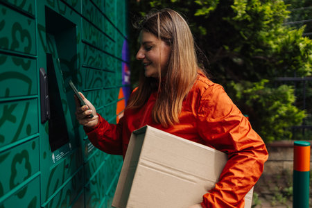 Caucasian woman picks up parcel from automatic post office machine, standing with phone near the self-service mail terminal. Parcel delivery machine.の写真素材