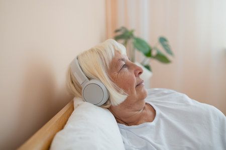 Elderly woman wearing headphones lying on bed using smartphone, embracing technology and enjoying a peaceful moment in her bedroom.の写真素材