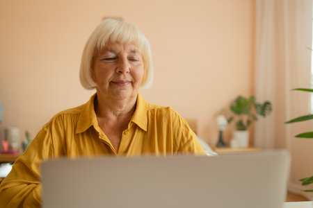 Happy senior woman looking at camera while working at home with laptop. Beautiful stylish elderly lady browsing site on cellphone. High quality photoの写真素材