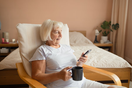 Caucasian 60s 70s happy woman using cell phone drinking coffee or tea relaxing at home. Smiling senior lady holding smartphone enjoying mobile shopping, checking social media messages, playing game or buying online.の写真素材