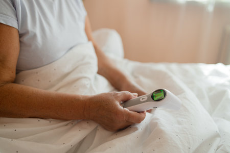 Caucasian 50s senior woman touching forehead after using thermometer to checking body temperature and take medicine lying under blanket on bed. Sick female with cold lying in bed with high fever in bedroom.の写真素材