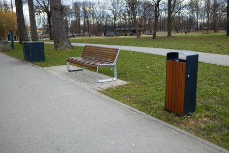 A well-maintained public park scene featuring a wooden bench, trash bins, and a green park setting. The path runs through the park with trees in the background, offering a peaceful atmosphereの写真素材