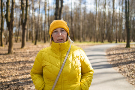 A thoughtful woman dressed in a yellow jacket and hat, standing on a park path surrounded by autumn trees and fallen leaves. The peaceful scene captures the beauty of the changing seasonsの写真素材