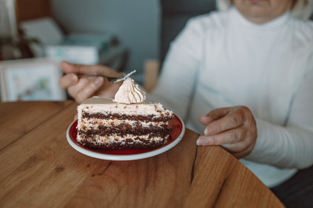 A close-up of a person eating a delicious slice of cake topped with cream, using a fork. The image captures a cozy, relaxed moment of enjoying a sweet dessert at a wooden table.の写真素材