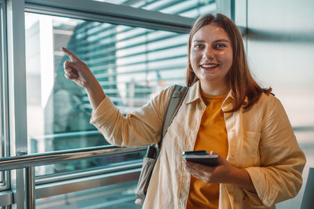 A cheerful young woman in a yellow shirt and jacket standing at the airport terminal. She is pointing at something with a smile, holding a smartphone in her hand. Ideal for travel, tourism, and lifestyle-related contentの写真素材