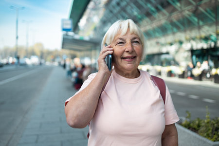 Senior woman is standing outside the airport terminal, talking on the phone with a smile. The bright...
