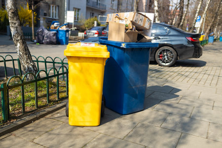 Two trash bins, one yellow and one blue, placed on the sidewalk. The bins are filled with cardboard boxes and waste, surrounded by greenery and urban buildings in the background. A clean street and vehicles are visible.の写真素材