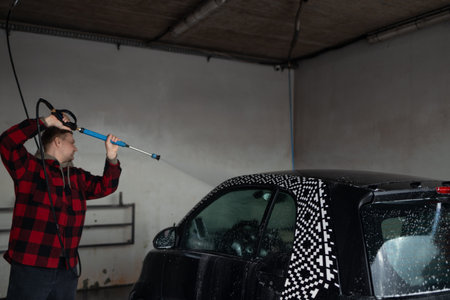 A man with a beard washes a gray car with a high-pressure washer in a car wash.の写真素材