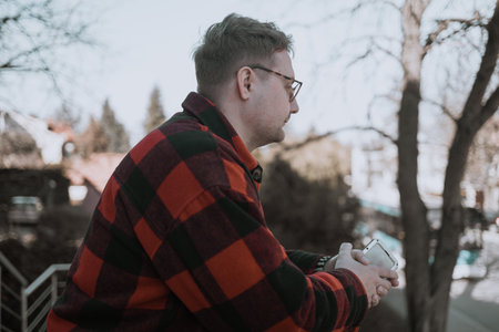 A young blond man wearing glasses, looking at his phone while walking down the street. A modern scene showcasing urban life, technology, and casual street styleの写真素材