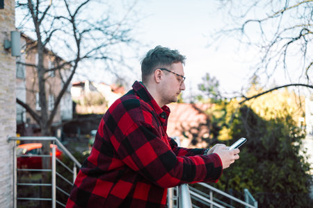 A young blond man wearing glasses, looking at his phone while walking down the street. A modern scene showcasing urban life, technology, and casual street styleの写真素材