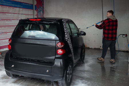 A man with a beard washes a gray car with a high-pressure washer in a car wash.の写真素材