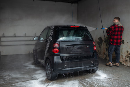 A man with a beard washes a gray car with a high-pressure washer in a car wash.の写真素材