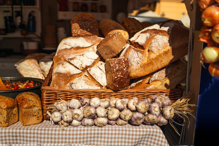 A variety of freshly baked loaves of bread are neatly arranged on a display table at a market. The loaves are golden-brown with crisp crusts and sit in a basket with a checkered cloth. Lights hang above, creating a warm atmosphereの写真素材
